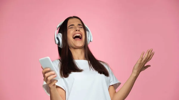 Брюнетка молода жінка в бездротових навушниках співає і тримає смартфон ізольовано на рожевому — стокове фото