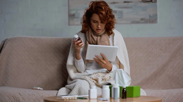 Kranke Frau benutzt digitale Tablette und hält Flasche mit Hustensaft in der Nähe von Medikamenten auf dem Tisch — Stockfoto