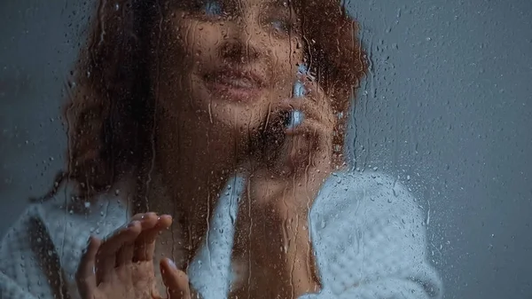 Cheerful and curly woman talking on smartphone behind window glass with raindrops — Photo de stock