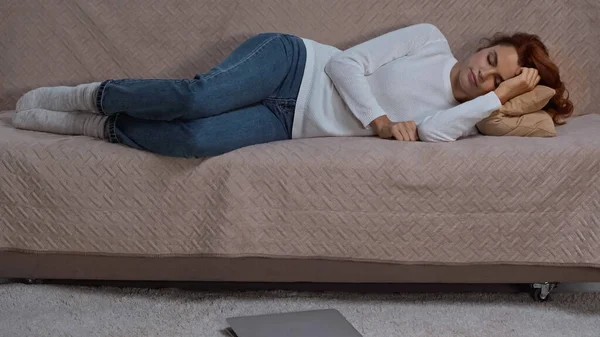 Redhead woman napping on couch near laptop in living room — Fotografia de Stock