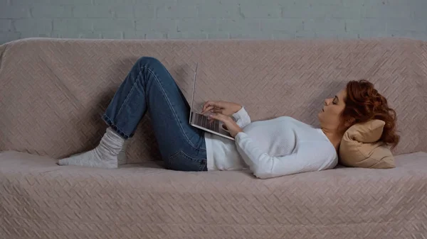 Side view of curly woman lying on couch and using laptop while working from home - foto de stock