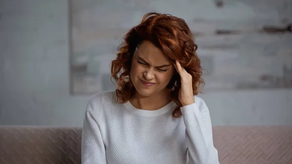 Ruiva mulher franzindo a testa enquanto sofre de dor de cabeça em casa — Fotografia de Stock