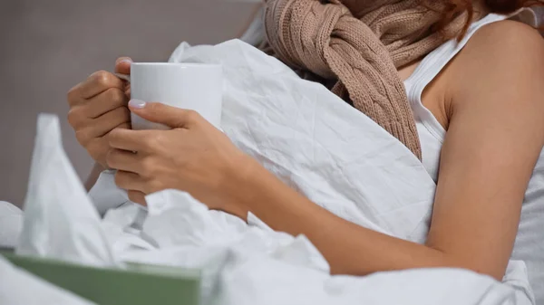 Cropped view of sick woman in scarf holding cup with beverage near blurred napkins — Photo de stock