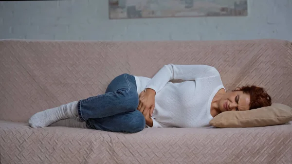 Young woman lying on sofa and suffering from cramps and having stomach ache — Fotografia de Stock