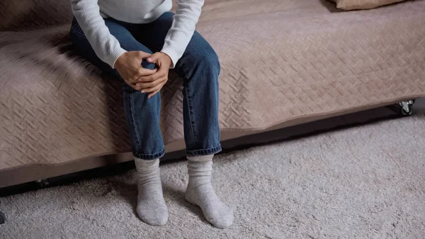 Cropped view of woman in jeans feeling knee pain while sitting on sofa in living room — Stock Photo
