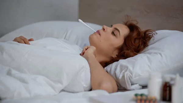 Redhead and sick woman measuring temperature with electronic thermometer and lying on bed — Foto stock