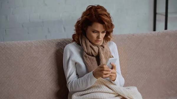 Sick and redhead woman in scarf sitting on couch and chatting on smartphone — Photo de stock