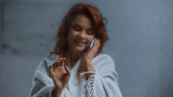 Smiling and curly woman talking on smartphone behind window glass with raindrops — Stockfoto
