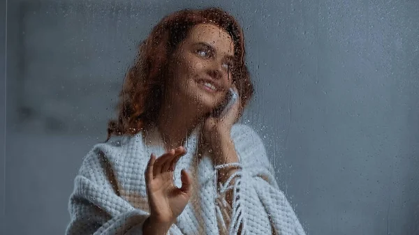 Positive and curly woman talking on smartphone behind window glass with raindrops — Foto stock