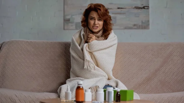 Sick and redhead woman talking at camera while sitting near medication on coffee table — Foto stock