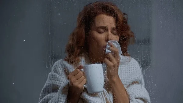 Sick woman holding cup with hot beverage and coughing behind window glass with raindrops — Stockfoto