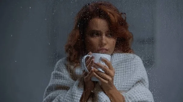 Sick woman holding cup with hot beverage behind window glass with raindrops — стоковое фото