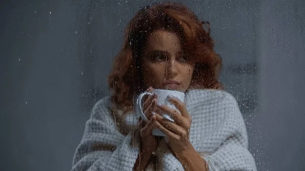 Sick woman holding cup with hot beverage behind window glass with raindrops — Stockfoto