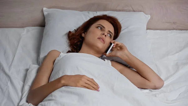 High angle view of displeased woman talking on smartphone while lying on bed — стоковое фото