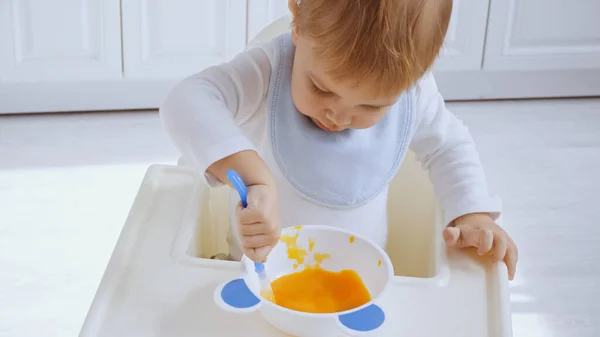 Cute toddler boy in bib sitting on baby chair and eating pumpkin puree — Stock Photo