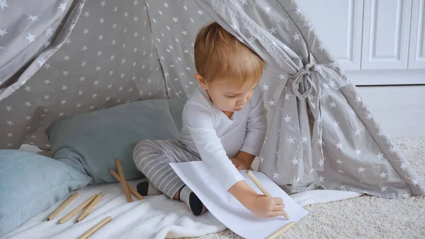 Concentrated toddler boy sitting on carpet in baby wigwam and drawing with color pencil on paper — Stockfoto