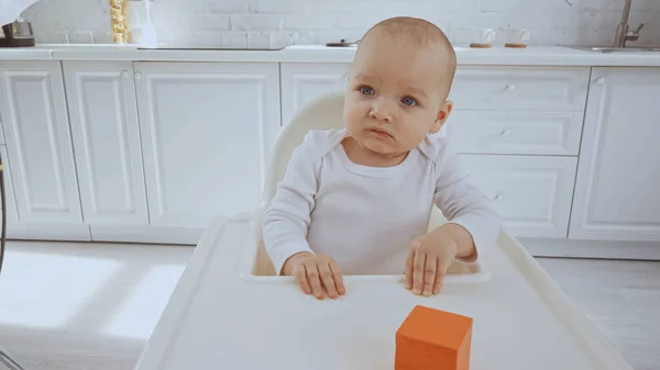 Confused baby girl sitting in feeding chair with orange wooden cube — Stock Photo