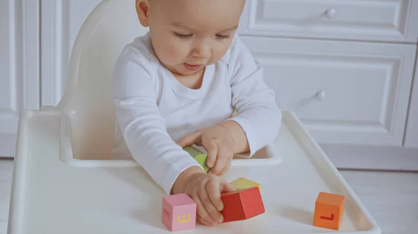 Focused baby girl sitting in feeding chair and playing with wooden cubes — Stock Photo