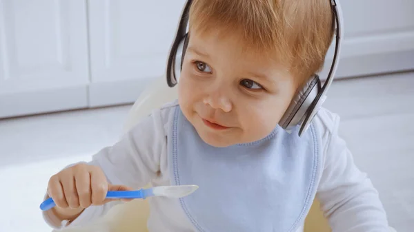 Happy toddler boy listening music in headphones while holding plastic spoon — Stock Photo
