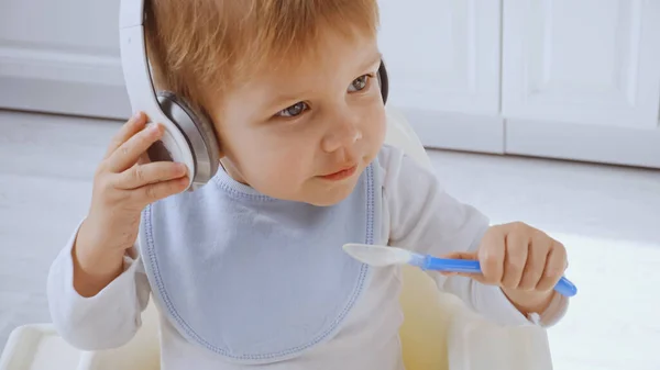 Cute toddler boy listening music in headphones while holding plastic spoon — Stock Photo