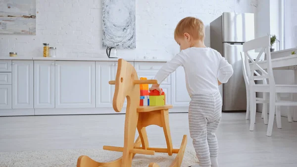 Toddler kid playing with building blocks on rocking horse in spacious apartment — стоковое фото