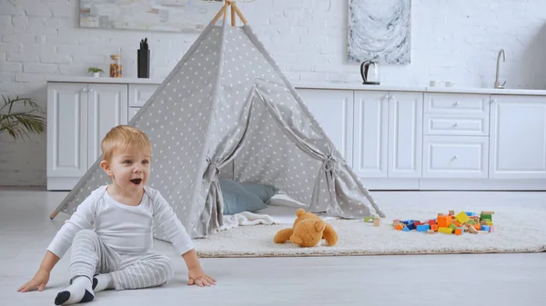 Excited toddler boy sitting near baby wigwam and toys on carpet — стоковое фото