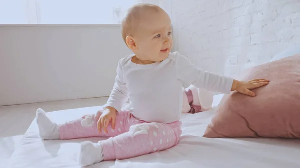 Happy toddler child sitting on bed and smiling while touching pink pillow — Stock Photo