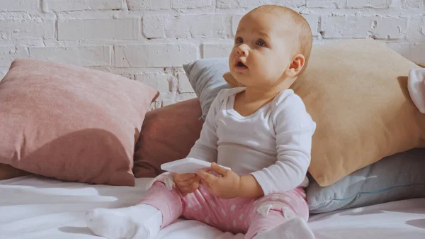 Surprised baby girl sitting on bed and holding smartphone around pillows — Foto stock