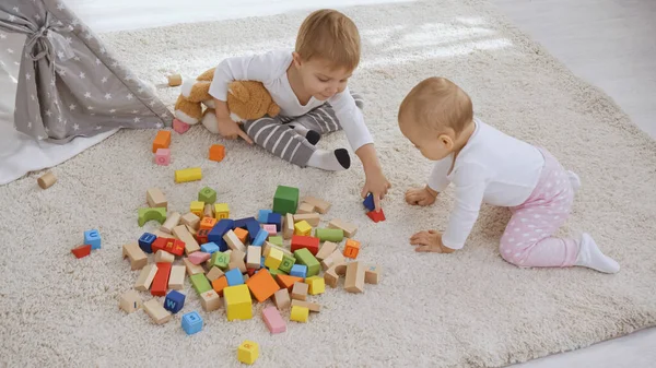 Toddler boy sitting on carpet with teddy bear near baby sister and playing with wooden blocks together — стоковое фото