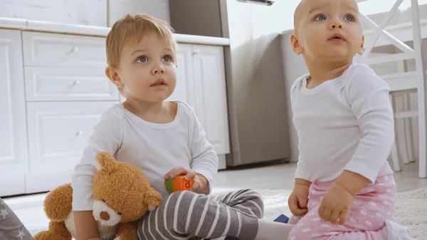 Toddler boy sitting on carpet with teddy bear near baby sister and looking away — Fotografia de Stock