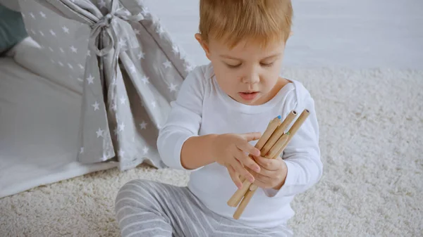 Concentrated toddler boy holding color pencil and sitting on carpet near baby wigwam - foto de stock