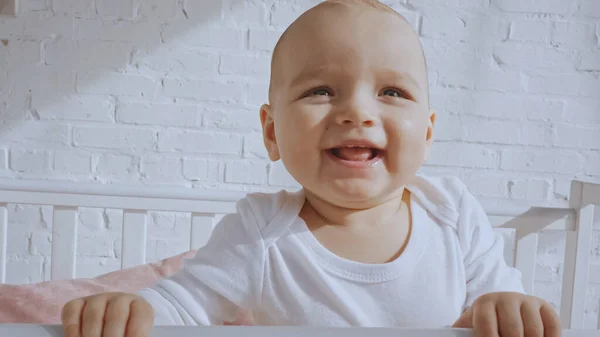 Cheerful baby standing in white baby crib while smiling at home — Stock Photo