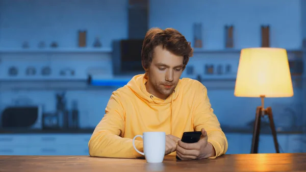 Зосереджений молодий чоловік використовує смартфон біля чашки чаю на дерев'яному столі — стокове фото