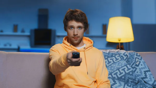 Young man in yellow hoodie sticking out tongue while clicking channels in living room — Photo de stock