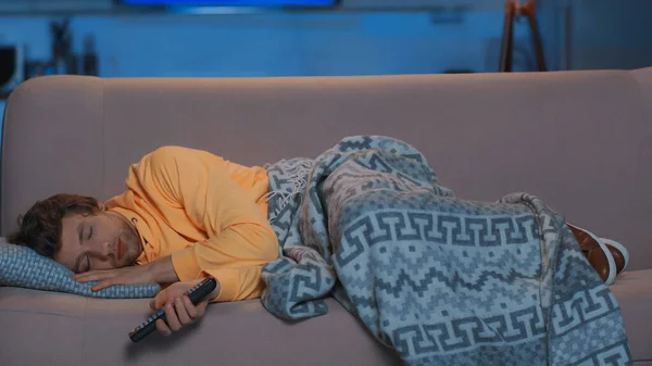 Young man in yellow hoodie holding remote controller and lying under blanket on sofa — стоковое фото