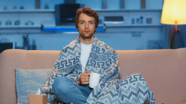 Young sick man sitting under blanket and holding cup of tea in living room — Stock Photo