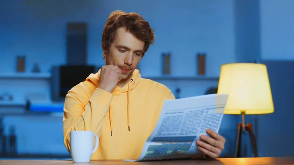 Young man in yellow hoodie reading newspaper near cup of tea at home — Stock Photo