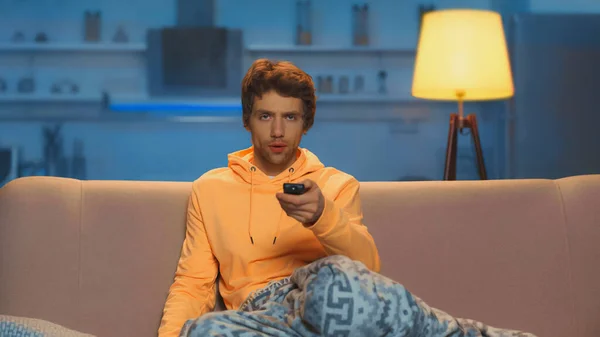 Young man in yellow hoodie holding remote controller and clicking channels — Stock Photo