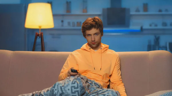 Skeptical young man in yellow hoodie holding remote controller and clicking channels — Foto stock