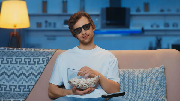 Pleased man in 3d glasses holding bowl with popcorn while watching movie in living room - foto de stock