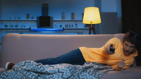 Focused young man in yellow hoodie near blanket and clicking channels at night — Stock Photo