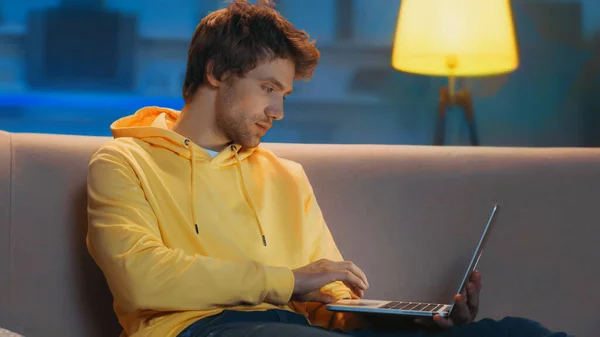 Young freelancer in yellow hoodie using laptop while working from home in evening — Stock Photo