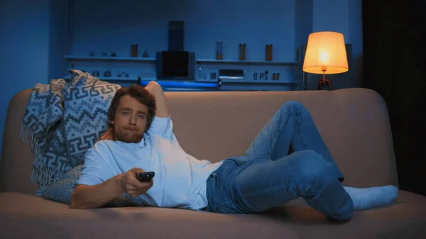 Bored young man lying on couch and watching tv in modern living room at night — Foto stock