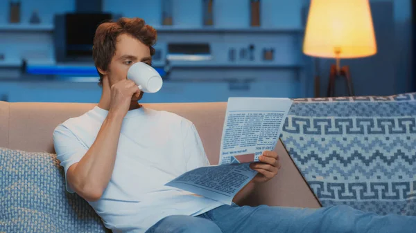 Молодий чоловік у білій футболці читає газету і п'є чай, відпочиваючи на дивані — стокове фото