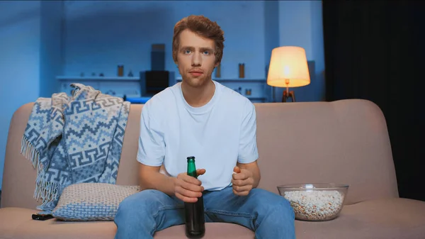 Focused man with bottle of beer sitting on couch near bowl of salty popcorn while watching championship — Photo de stock