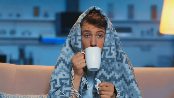 Sick man with blue eyes drinking hot beverage while sitting under blanket in living room - foto de stock