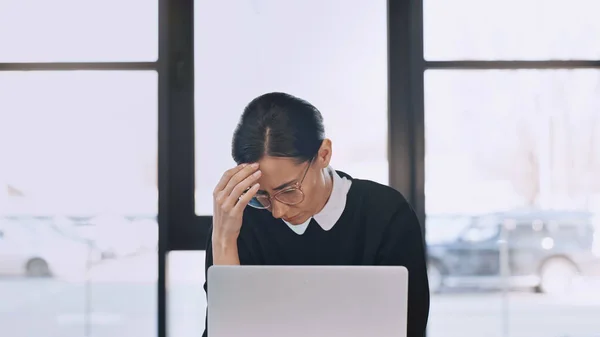 Exhausted businesswoman in eyeglasses touching forehead while suffering from headache near laptop — стоковое фото