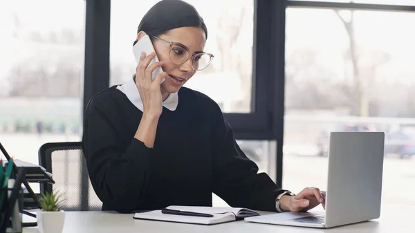 Brunette businesswoman in eyeglasses using laptop and talking on smartphone in office — стоковое фото