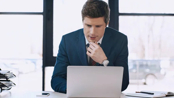 Pensive businessman touching chin and looking at laptop in office — Stock Photo