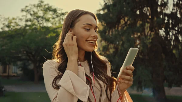 Positive woman using earphones and smartphone in blurred summer park — Stock Photo